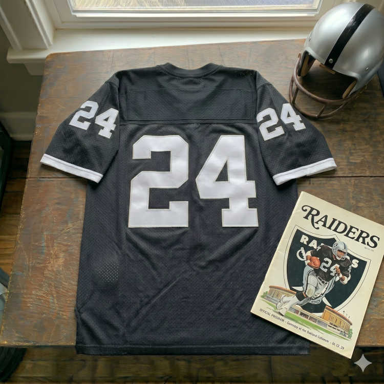 Vintage Oakland Raiders #24 football jersey. Black mesh with solid white block numbers and clean white sleeve cuffs. Features Raiders game program at Oakland Coliseum and silver helmet for historical AFL-NFL merger research and Pigskin Dispatch archives.