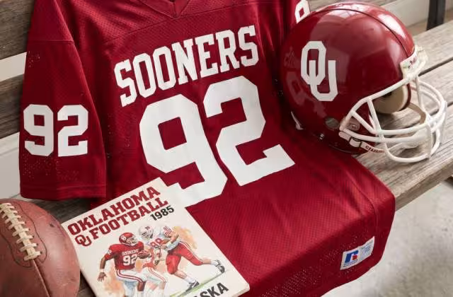 Gemini AI-created image of a 1985 Oklahoma Sooners football jersey and helmet on a locker room bench. The jersey features the requested number 92 without a player's name and is accompanied by a period-correct helmet and game program.