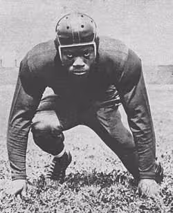 Vintage black and white image of an intimidating Bill Willis (1921–2007) strikes an intimidating middle guard pose as part of the Ohio State Buckeyes football team in 1943. Photographer uncredited. Published in Francis Powers, "The West," in Jack Byrne and Malcom Reiss (eds.), 1943 Illustrated Football Annual. New York: Fiction House Publishers, 1943; p. 49.