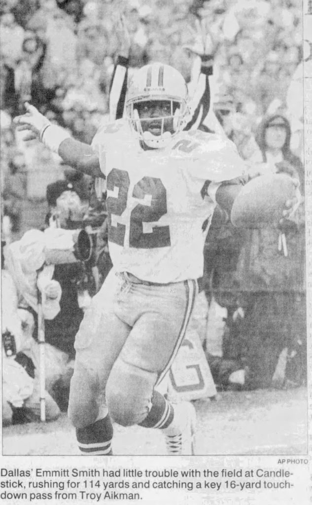 A black-and-white AP Photo clipping of Dallas Cowboys running back Emmitt Smith (#22) celebrating a touchdown. He is shown with his arms outstretched, holding the football in his left hand, after a play at Candlestick Park. The caption reads: 'Dallas' Emmitt Smith had little trouble with the field at Candlestick, rushing for 114 yards and catching a key 16-yard touchdown pass from <a rel=