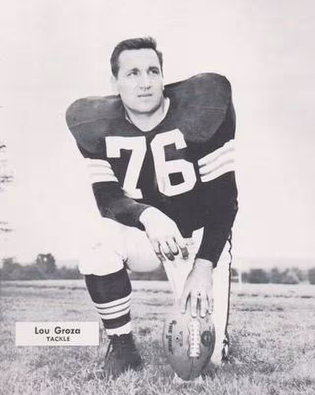 b/w photo promo of Lou Groza in his dark colored number 76 Browns jersey kneeling on one knee with a teed football