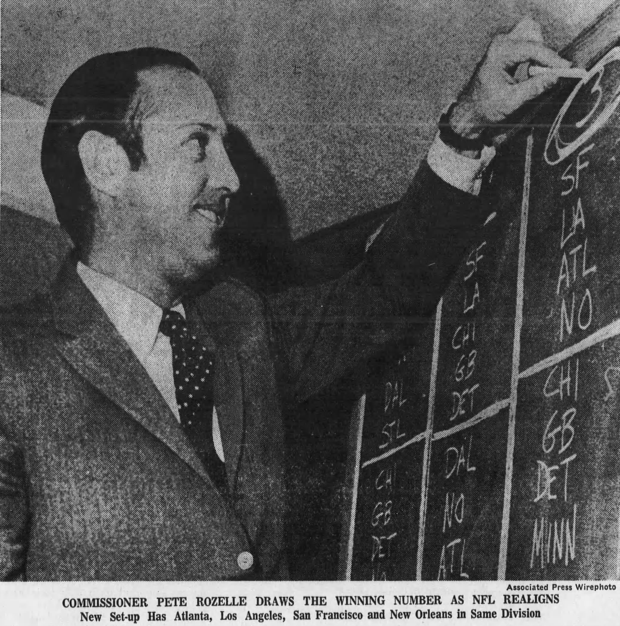 vintage black and white newspaper image of NFL Commissioner Pete Rozelles at a blackboard, smiling as he chalks something up.