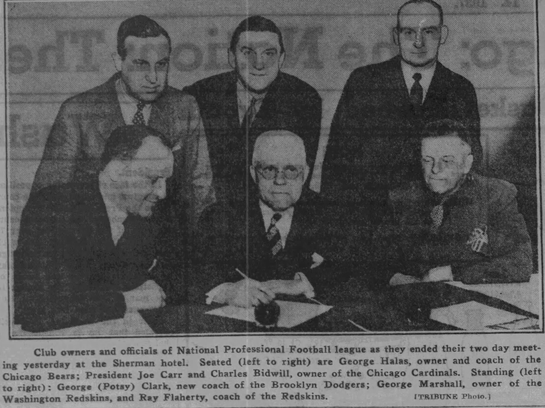 black and white image of the 1937 NFL owners meeting with 6 men: George Halas, Joe Carr, Charles Bidwell, Potsy Clark, George Marshall, and Ray Flaherty posing in front of a table filled with documents to sign