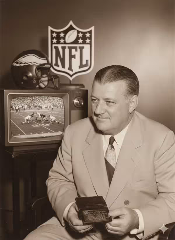 AI generated imposing of Black and white image of a cropped Bert Bell sitting at a table with a TV set, a n Eagles helmet and the NFL shield in the background