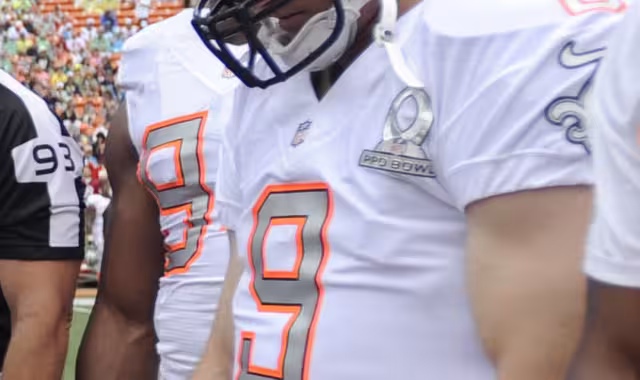 color image of NO Saints QB Drew Brees in a white 2014 NFC Pro Bowl Jersey and Saint helmet at midfield with teammates and an official