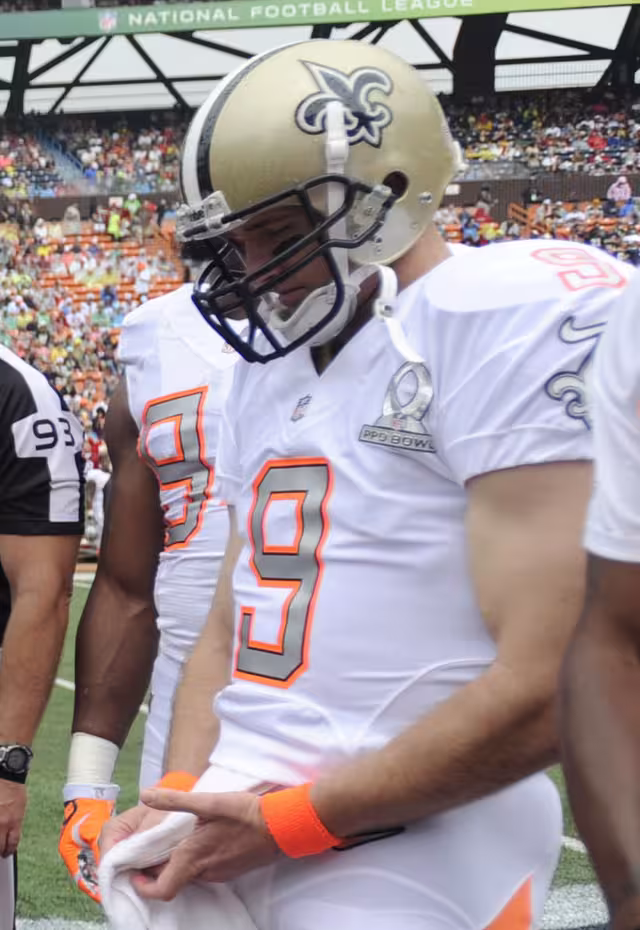 color image of NO Saints QB Drew Brees in a white 2014 NFC Pro Bowl Jersey and Saint helmet at midfield with teammates and an official