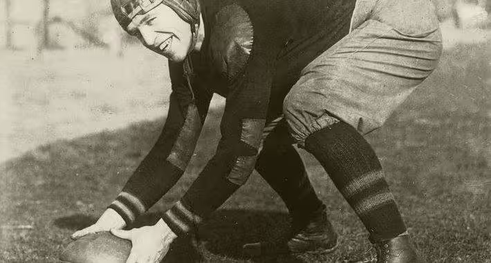black and white full image of John McEwan in full gridiron gear and lether helmet posing as if ready to snap a football