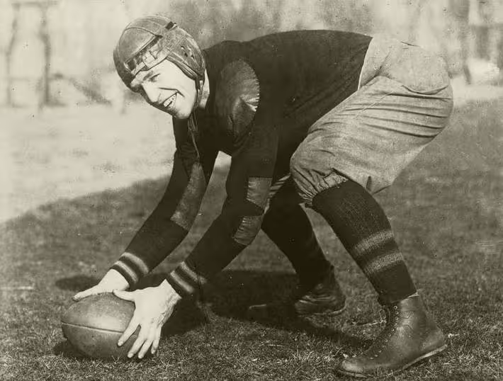 black and white full image of John McEwan in full gridiron gear and lether helmet posing as if ready to snap a football