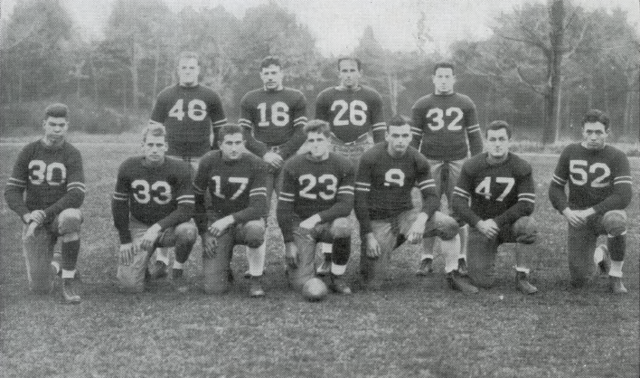 A black-and-white team photograph of eleven members of the 1935 New Hampshire Wildcats football team. The players are posed in two rows on a grass field—four standing in the back and seven kneeling in the front. They wear dark, heavyweight jerseys with large white block numbers and double stripes on the sleeves. A vintage leather football rests in front of the center kneeling player. The background is a dense line of autumn trees.