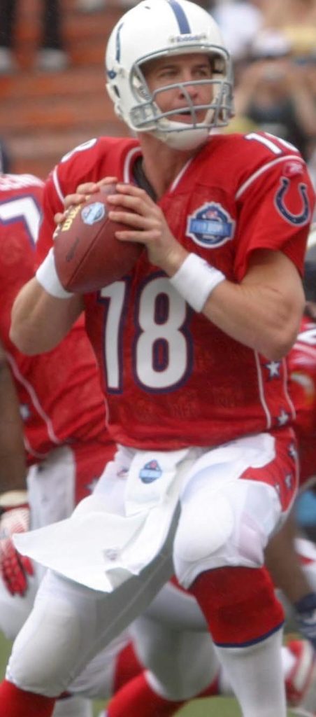 A color action photograph of quarterback Peyton Manning during an NFL Pro Bowl game. Manning is wearing the AFC's red jersey with his iconic number '18' in white, paired with white pants and a white Indianapolis Colts helmet featuring the blue horseshoe logo. He is shown in a classic passing pocket stance, clutching the football with both hands near his chest. The Pro Bowl patch is visible on the right chest of his jersey.