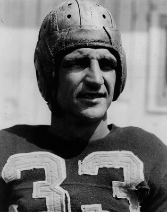 A vintage black-and-white close-up portrait of Hall of Fame quarterback Sammy Baugh. He is wearing a dark jersey with the number '33' in large, light-colored block digits and a classic leather suspension helmet with visible ventilation holes and protective ridges. He has a rugged, focused expression, looking slightly off-camera.
