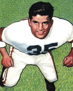 A color-tinted photograph of Alex Agase from a 1950 Bowman football card. Agase is shown in a three-quarter view from a high angle, crouching in a football stance on a vibrant green grass field. He is wearing a white Cleveland Browns jersey with his number '35' in dark block numerals and white pants with a vertical orange and brown stripe. His expression is determined as he looks upward, with both hands clenched in fists.