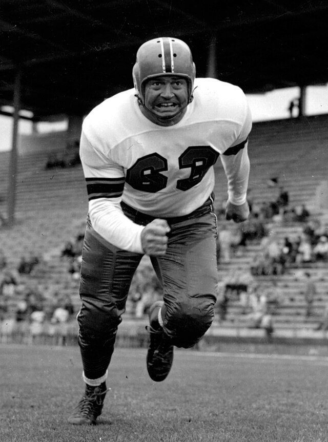 A black-and-white action photograph of B.C. Lions defensive tackle Arnie Weinmeister in 1954. He is captured mid-stride in a low, powerful charging stance on a grass field. He is wearing a white jersey with the number '63' in dark block digits and a dark-striped helmet. His expression is intense and focused, with gritted teeth. Empty stadium bleachers are visible in the background.