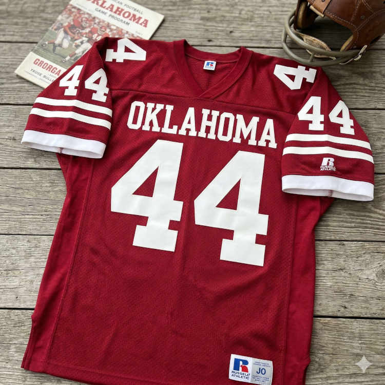 A flat-lay photograph of a crimson Oklahoma Sooners football jersey on a weathered grey wood surface. The mesh jersey features 'OKLAHOMA' and the number '44' in bold white block lettering. In the background, a vintage Oklahoma game program and a classic leather helmet with a grey facemask are visible