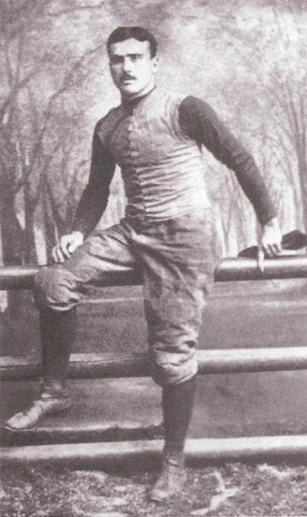 A sepia-toned archival photograph of Yale football legend Thomas 'Bum' McClung from the late 19th century. He is shown standing outdoors, leaning one leg against a wooden rail fence. He wears a traditional canvas sleeveless vest over a dark, long-sleeved jersey, paired with padded knickers and high-top lace-up leather boots. His expression is stoic, capturing the rugged aesthetic of the 'scrum' era of football.
