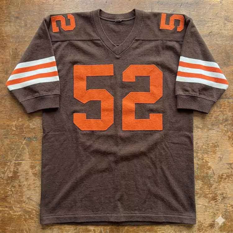 A flat-lay photograph of a vintage dark brown football jersey on a rustic wooden surface. The jersey features the number '52' in bold orange block lettering on the chest and shoulders. The sleeves are accented with three horizontal stripes in a white-orange-white pattern, and the jersey has a ribbed V-neck collar.