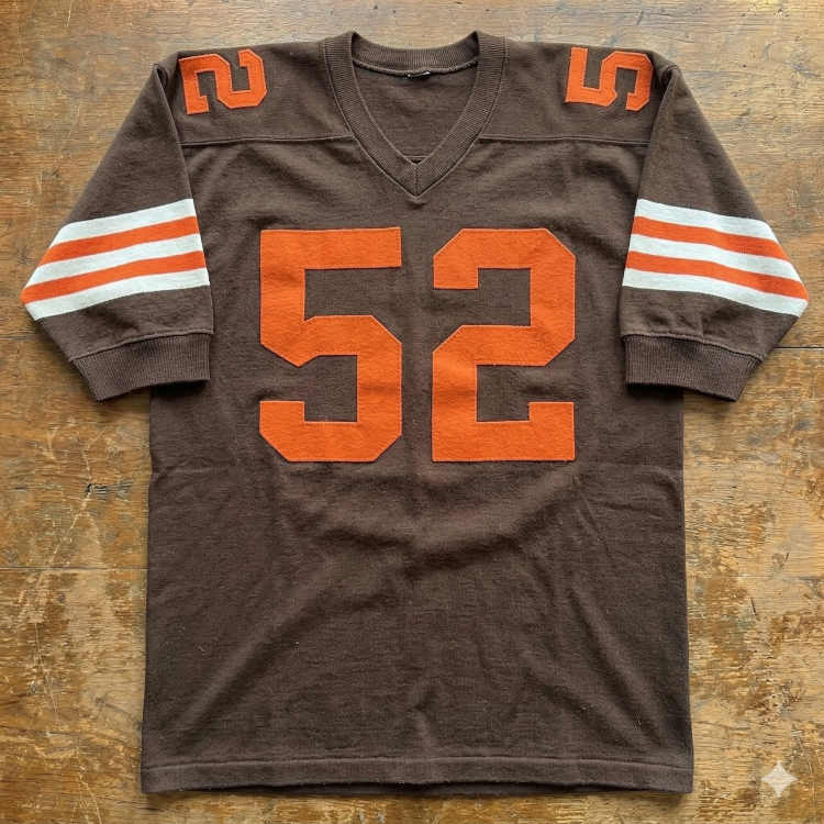 A flat-lay photograph of a vintage dark brown football jersey on a rustic wooden surface. The jersey features the number '52' in bold orange block lettering on the chest and shoulders. The sleeves are accented with three horizontal stripes in a white-orange-white pattern, and the jersey has a ribbed V-neck collar.
