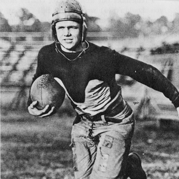 A black-and-white archival photograph of Georgia Tech's Bill Flowers in a dynamic running pose. He is shown mid-stride on a grass field, clutching an early, more rounded football in his right arm. He wears a dark, long-sleeved jersey with a light-colored triangular 'V-neck' panel on the chest, a soft leather helmet with a small brim, and padded, lace-up trousers. The background shows blurred stadium bleachers on a sunny day.