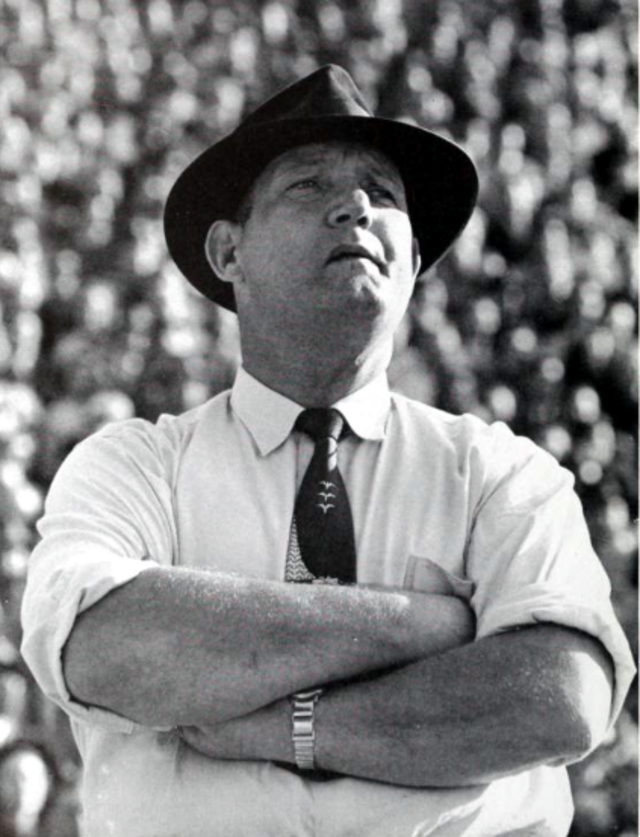 A black-and-white archival photograph of Clemson head coach Frank Howard from the 1956 'Taps' yearbook. Howard is shown from the waist up, arms crossed, wearing a light-colored button-down shirt, a dark patterned necktie, and a classic fedora. He is gazing upward with a focused expression. A blurred crowd in the stadium bleachers provides the background, capturing a candid moment on the sidelines.
