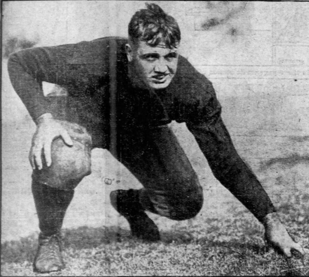 A black-and-white action photograph of Frank Wickhorst from circa 1926. He is captured in a powerful three-point stance on a grass field, wearing a dark, long-sleeved football jersey and padded trousers. He has a determined expression, and the grainy texture of the newsprint reflects the era of his All-American career at the United States Naval Academy."