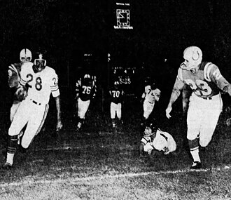 A black-and-white action photograph from a 1958 NFL game. Chicago Bears running back Willie Galimore (#28) is shown sprinting toward the left with the football tucked in his arm. Baltimore Colts defensive back Art DeCarlo (#23) is in pursuit from the right. Several other players are visible in the background, along with a stadium scoreboard showing '5:05' remaining in the quarter. The scene is illuminated by stadium lights for a night game.