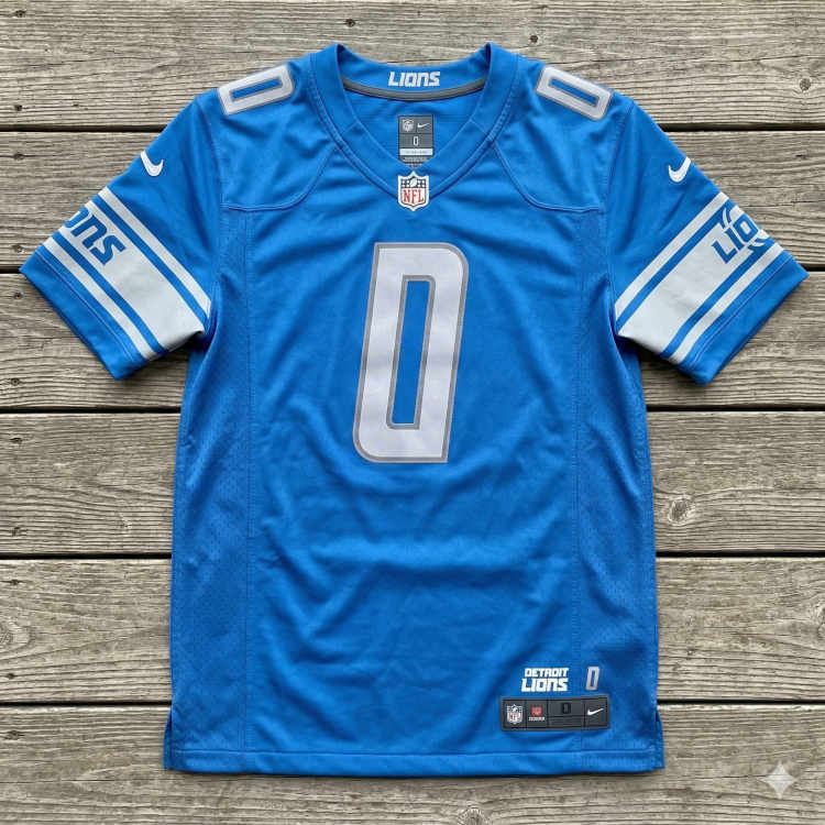 A flat-lay photograph of a Honolulu blue Detroit Lions jersey displayed on a grey wooden deck. The jersey features the number '0' in a modern silver-and-white font on the chest and shoulders. The sleeves are accented with wide silver bands containing the 'LIONS' wordmark and team logo. A silver Nike 'Swoosh' is visible on the shoulders, and an NFL Shield sits at the base of the V-neck collar.