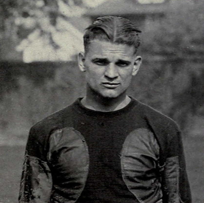 A black-and-white portrait of Michigan football legend Harry Kipke from 1922. He is shown from the chest up, sporting a classic 1920s side-parted hairstyle. He wears a dark, heavyweight wool jersey featuring two large, circular leather friction patches on the chest—a common design element of the era to assist in ball security. The fabric shows authentic signs of wear and field dirt, particularly on the sleeves.