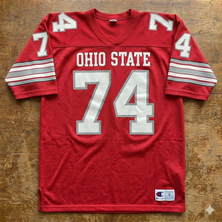 A flat-lay photograph of a scarlet red mesh football jersey displayed on a weathered wooden surface. The jersey features the words 'OHIO STATE' in white block letters above a large number '74' in silver digits with a white outline. The sleeves include horizontal grey, white, and red stripes with smaller numbers on the shoulders. A white Champion jock tag is sewn onto the lower right hem.