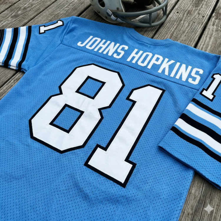 A flat-lay photograph of a light blue Johns Hopkins football jersey on a weathered wood surface. The mesh jersey features the school name 'JOHNS HOPKINS' and the number '81' in bold white block lettering with dark outlines. The top of a grey football helmet with a dark facemask is visible at the top edge of the frame.