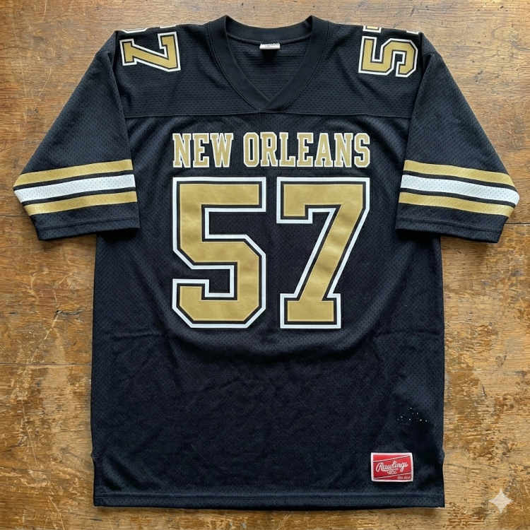 A flat-lay photograph of a black mesh football jersey displayed on a weathered wooden surface. The jersey features the words 'NEW ORLEANS' arched in gold block letters above a large number '57' in gold digits with a white-and-black outline. The sleeves include horizontal gold-and-white stripes and smaller numbers on the shoulders. A vintage red Rawlings jock tag is sewn onto the lower right hem.