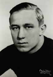 A black-and-white studio headshot of Jay Berwanger, the 1935 Heisman Trophy winner. He is shown from the shoulders up, wearing a dark, high-neck athletic sweater. He has short, light-colored hair neatly parted on the side and a serious, steady expression. A photographer's signature, 'Paul Stone-Raymor,' is visible in the lower right corner.