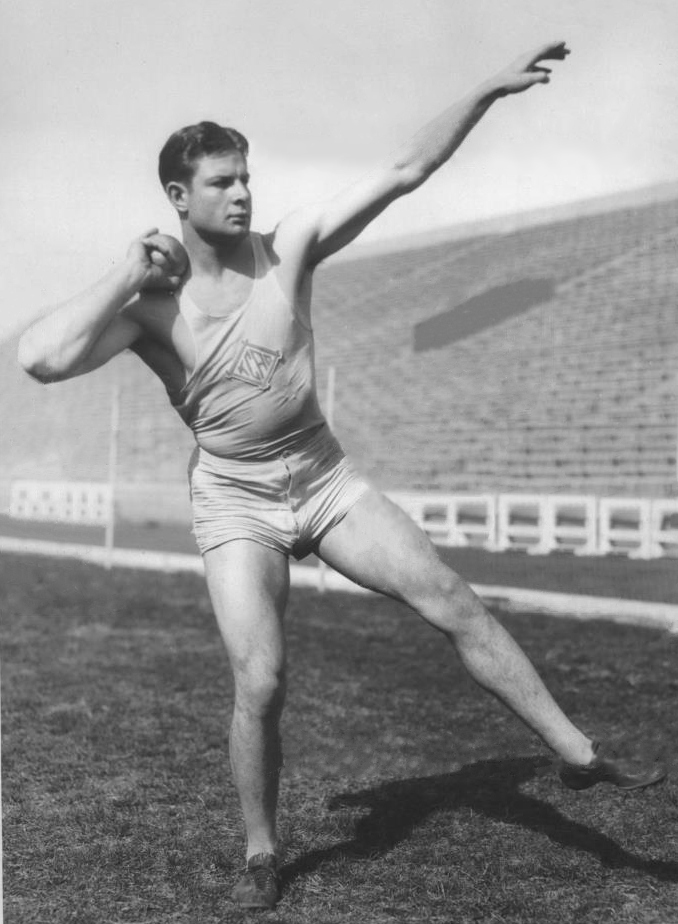 A black-and-white archival photograph of Jim Bausch from 1931, captured in the middle of a shot put throw. He is shown in a dynamic athletic pose on a grass field, with one arm extended for balance and the other holding the shot near his neck. He wears a light-colored 'KCAC' (Kansas City Athletic Club) tank top and short athletic trunks. The background features the empty, sun-drenched bleachers of a large stadium.