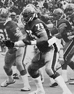 A black-and-white action photograph of offensive tackle Jimbo Covert playing for the University of Pittsburgh in 1980. Covert, wearing his #75 jersey and a dark helmet with a center stripe, is captured in a powerful three-point stance on the line of scrimmage. He is wearing white mid-calf socks and dark gloves, with a crowd visible in the background.