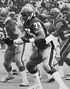A black-and-white action photograph of offensive tackle Jimbo Covert playing for the University of Pittsburgh in 1980. Covert, wearing his #75 jersey and a dark helmet with a center stripe, is captured in a powerful three-point stance on the line of scrimmage. He is wearing white mid-calf socks and dark gloves, with a crowd visible in the background.