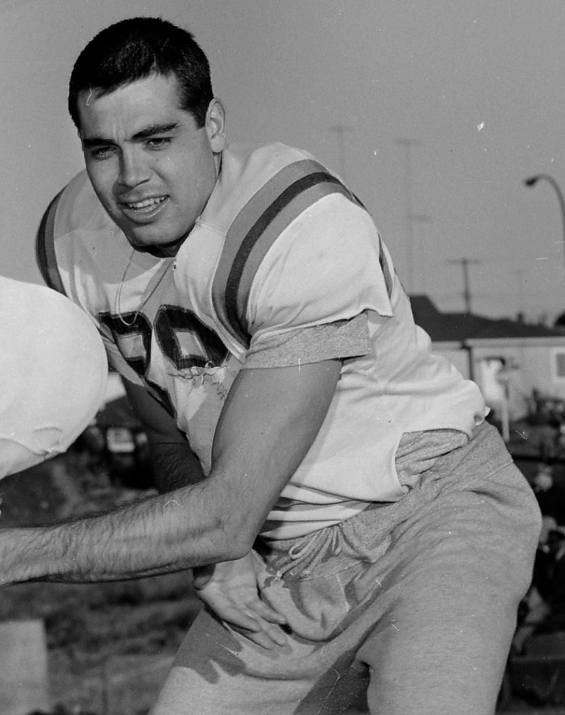 A black-and-white candid action photograph of quarterback Joe Kapp. He is captured mid-play in a crouched athletic stance, looking focused and determined. He is wearing a light-colored jersey with dark shoulder stripes and large digits on the chest, paired with drawstring training pants. The image highlights his rugged, unmasked presence on the field.
