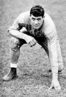 A vintage black-and-white photograph of Joe Stydahar during his tenure at West Virginia University (circa 1933–1935). He is shown in a three-point football stance on a grass field, wearing a dark long-sleeved jersey, padded trousers, and high-top leather cleats. He has a focused expression looking directly at the camera.