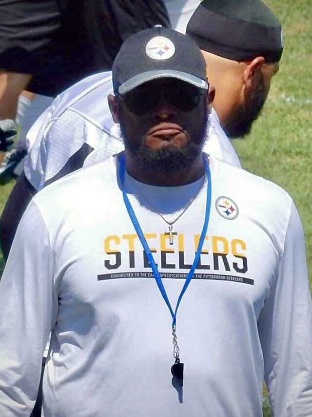 A close-up photograph of Pittsburgh Steelers head coach Mike Tomlin during the 2016 training camp. He is wearing a white long-sleeved Steelers shirt, a black team cap with the Steelers logo, and dark sunglasses. A whistle hangs around his neck on a blue lanyard, and he is looking toward the camera with a focused expression.