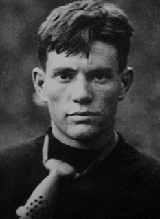 A black-and-white close-up portrait of Nathan Dougherty in his University of Tennessee football attire. He is shown from the chest up, wearing a dark, high-neck jersey. Hanging around his neck is a vintage leather 'nose mask' or nose guard, which features several ventilation holes. He has short, dark hair and a focused, intense gaze.