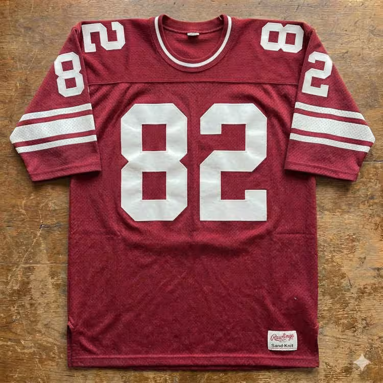 A flat-lay photograph of a maroon mesh football jersey displayed on a rustic wood-grain surface. The jersey features the number '82' in large white block digits on the chest and shoulders. The sleeves are accented with two white horizontal stripes, and a vintage Rawlings Sand-Knit jock tag is visible at the lower right hem.
