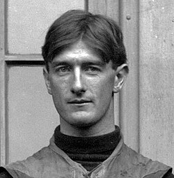 A vintage black-and-white close-up portrait of Pat O'Dea, the 'Kangaroo Kicker.' The image captures O'Dea from the shoulders up, wearing a dark turtleneck sweater under his canvas football vest. He has a steady, direct gaze and short, middle-parted hair, set against the backdrop of a wooden paneled door.
