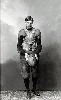 A full-length vintage black-and-white studio portrait of Hall of Fame kicker Pat O'Dea from the University of Wisconsin. He is standing in full period-accurate football gear, including a lace-up canvas vest, padded trousers, and high-top cleats. He is holding a melon-shaped football in both hands, positioned as if preparing for a drop kick against a clouded studio backdrop.