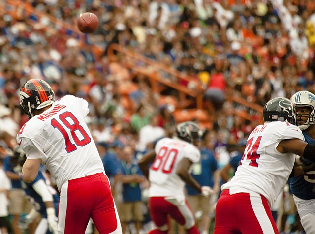 A color action photograph from the 2013 NFL Pro Bowl. Quarterback Peyton Manning (#18) is shown from behind, releasing a pass. He is wearing the AFC's white jersey with red numbers and red trousers, but notably, he is sporting his dark Denver Broncos helmet with the orange mane logo. In the foreground, Houston Texans offensive lineman Chris Myers (#55) and Wade Smith (#74) provide protection against a Detroit Lions defender.