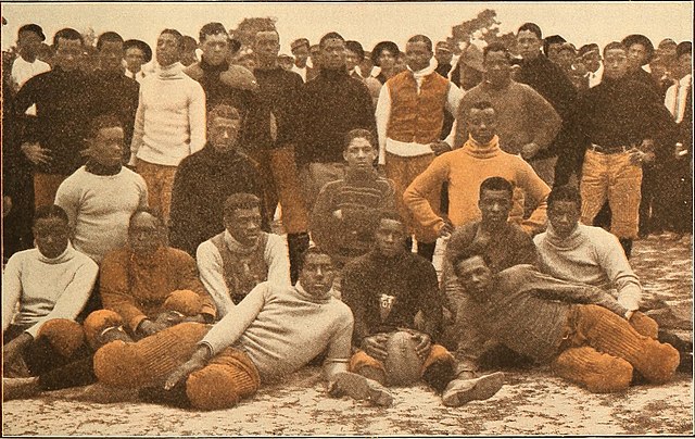 A sepia-toned group portrait of a large football squad from 1908. The team is composed of African American athletes posing on a dirt field. They are wearing a variety of early gridiron apparel, including heavy turtleneck sweaters, padded canvas trousers, and dark long-sleeved jerseys. A player in the front row sits cross-legged holding a lace-up leather football, while others are seen standing or reclining in a classic team-photo arrangement.