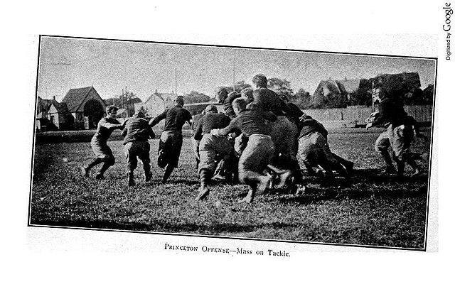 A grainy, black-and-white historical photograph titled 'Princeton Offense—Mass on Tackle.' The image captures a dense group of players in dark jerseys and light-colored quilted pants engaged in a 'mass play' on a grass field. Several players are bunched together to push through the defensive line, while others are seen running toward the scrum in a wide-open field setting with houses visible in the background.
