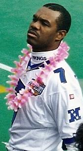 A color photograph of quarterback Randall Cunningham at the 1999 NFL Pro Bowl in Honolulu, Hawaii. He is wearing a white NFC Pro Bowl jersey with purple 'VIKINGS' branding on the collar and his number '7' on the sleeve. A pink flower lei is draped around his neck, and he has a focused, upward-looking expression. The bright green turf of Aloha Stadium is visible in the background.