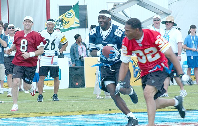A vibrant color photograph of several NFL legends playing flag football. On the left, Steve Young (#8) in a maroon jersey is running toward the play. In the center, Michael Irvin (#88) in a navy Dallas Cowboys jersey is sprinting with the football. On the right, Marcus Allen (#32) in a red Kansas City Chiefs jersey is in pursuit. In the background, Rod Woodson (#26) in a white Pittsburgh Steelers jersey looks on. The players are wearing blue flag-football belts over athletic shorts on a grass field."