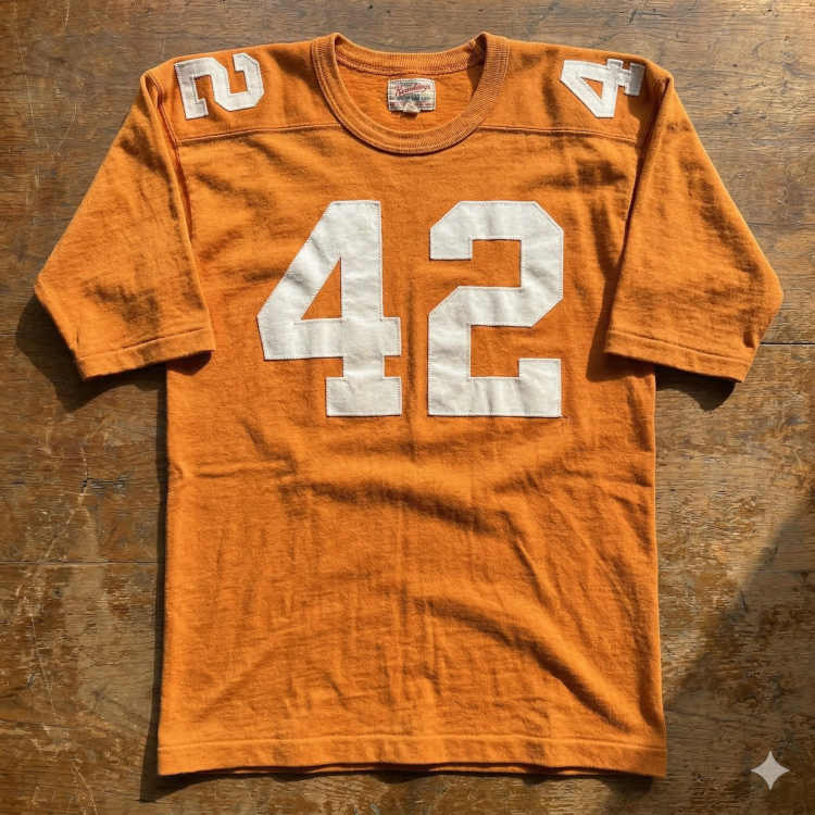 A flat-lay photograph of a vintage orange football jersey on a weathered wooden surface. The jersey features the number '42' in large white block digits on the chest and smaller digits on the shoulders. It is made of a heavy cotton or wool-blend fabric with a ribbed crew neck collar and a vintage manufacturer's tag visible inside the neck.