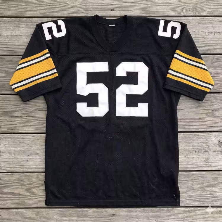 A flat-lay photograph of a black mesh football jersey displayed on a grey wooden deck. The jersey features the number '52' in large white block digits on the chest and shoulders. The sleeves are accented with broad gold horizontal bands, each containing two thin black and white stripes.