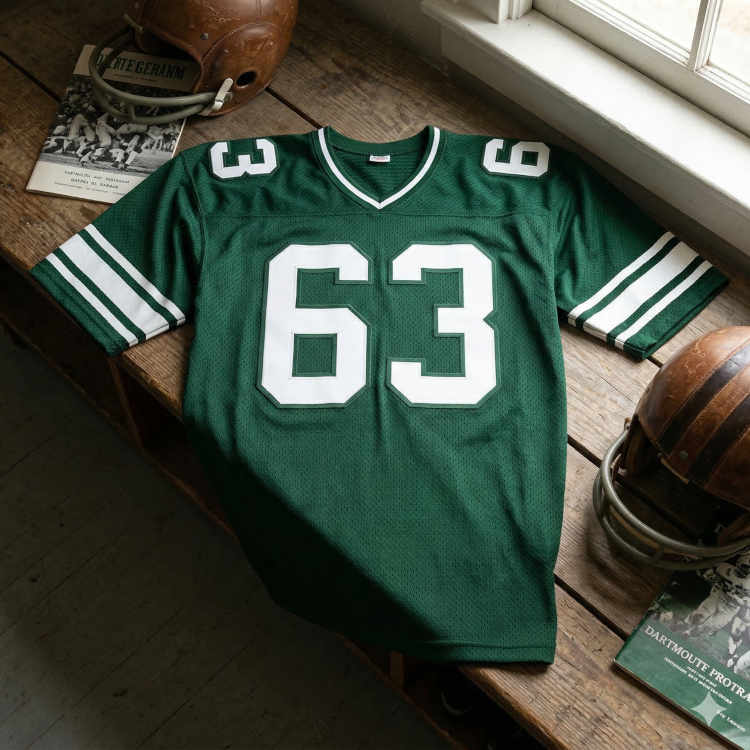 A photograph of a forest green mesh football jersey displayed on a weathered wooden locker bench by a window. The jersey features the number '63' in large white block digits on the chest and shoulders, with white trim on the V-neck and triple white stripes on the sleeves. Accompanying the jersey are two vintage leather helmets with faceguards and several 'Dartmouth' game programs.