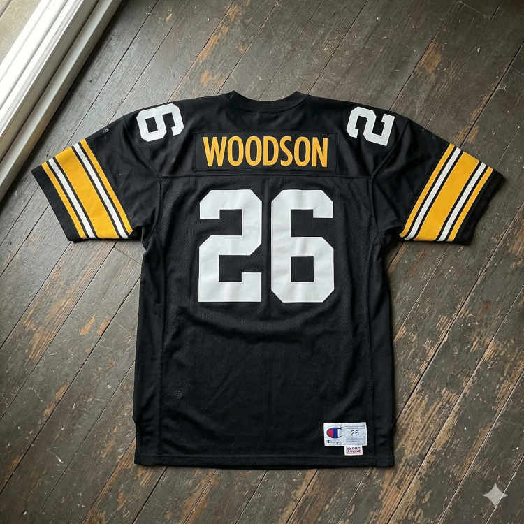 A flat-lay photograph showing the back of a black mesh Pittsburgh football jersey on a dark wood-plank floor. The name 'WOODSON' is printed in gold block letters on a black nameplate above the number '26' in large white digits. The sleeves feature broad gold bands with thin black and white stripes, and a Champion 'Pro Line' jock tag is visible at the bottom right.